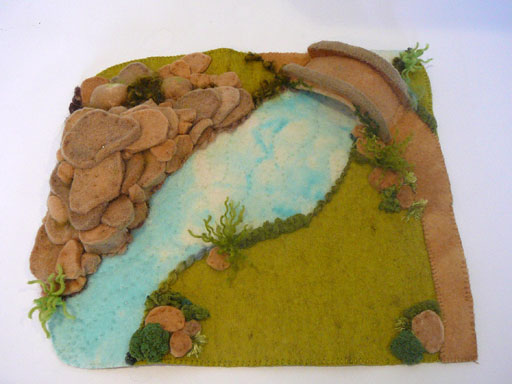 Story mat made from felt showing a birdge corssing a river,on oneside rocky terrain,the other a lush grass. 